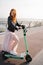 Beautiful happy young ginger girl using electric scooter in fashinable long pink dress black lether jacket