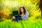 Beautiful happy young girl sits in a park on a glade with a laptop in green grass. sunny day