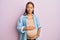 Beautiful hispanic woman expecting a baby, touching pregnant belly making fish face with mouth and squinting eyes, crazy and