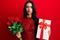 Beautiful hispanic woman holding anniversary gift and roses bouquet skeptic and nervous, frowning upset because of problem