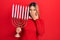 Beautiful hispanic woman holding menorah hanukkah jewish candle covering mouth with hand, shocked and afraid for mistake