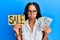 Beautiful hispanic woman holding sale banner and dollars puffing cheeks with funny face