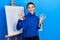 Beautiful hispanic woman with short hair holding manikin close to easel stand doing ok sign with fingers, smiling friendly