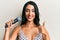 Beautiful hispanic woman singing song using microphone smiling happy and positive, thumb up doing excellent and approval sign