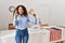 Beautiful hispanic woman standing by at political campaign by voting ballot gesturing finger crossed smiling with hope and eyes