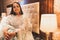 A beautiful Indian bride in traditional wear waiting in Hotel room next to lamp. A smiling girl wearing jewellery and designer