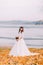 Beautiful innocent thoughtful bride in gorgeous white dress stands on fallen leaves at riverside