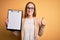 Beautiful inspector woman wearing glasses holding checklist clipboard over yellow background happy with big smile doing ok sign,