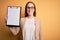 Beautiful inspector woman wearing glasses holding checklist clipboard over yellow background with a happy face standing and