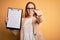 Beautiful inspector woman wearing glasses holding checklist clipboard over yellow background pointing with finger to the camera