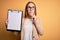 Beautiful inspector woman wearing glasses holding checklist clipboard over yellow background serious face thinking about question,