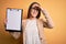 Beautiful inspector woman wearing glasses holding checklist clipboard over yellow background stressed with hand on head, shocked