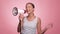 A beautiful joyful girl in a striped T-shirt shouts into the loudspeaker on isolated pink background