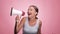 A beautiful joyful girl in a striped T-shirt shouts into the loudspeaker on isolated pink background