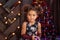 Beautiful kid girl 5-6 year old wearing stylish dress sitting in armchair over Christmas tree in room. Looking at camera. Holiday