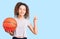 Beautiful kid girl with curly hair holding basketball ball surprised with an idea or question pointing finger with happy face,