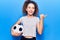 Beautiful kid girl with curly hair holding soccer ball pointing thumb up to the side smiling happy with open mouth