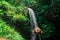 Beautiful lady in straw hat from back in greenery. Girl walks in the jungle and llooks at a strong and rapid waterfall. Adventure