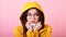 Beautiful lady in a yellow cloak with a hood on her head with glasses, snuggles into her hands because of the cold on a pink