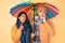 Beautiful latin young couple under colorful umbrella serious face thinking about question with hand on chin, thoughtful about