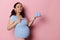 Beautiful Latina pregnant woman pointing finger aside cute blue baby booties on her hand, isolated on pink background.