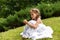 A beautiful little girl in a white long dress sitting on the bright green grass and looking