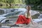 Beautiful long-haired girl in a red skirt sitting on a rock on a background of mountain river cascade. Summer evening. Tranquility