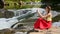 Beautiful long-haired girl in a red skirt sitting on a rock and making selfie on the background of a mountain river cascade. Summe