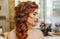 Beautiful, with long, red-haired hairy girl, hairdresser weaves a French braid, in a beauty salon.