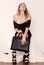 Beautiful luxurious young woman in a black dress sits on a white fur cover in black fashionable sandals and a handbag