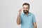 Beautiful man showing small thing while smiling and standing near white wall. Handsome bearded person in a blue shirt