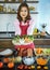 Beautiful mature adult Asian female housewife in apron standing in kitchen with various kinds and colorful vegetables and fruit