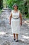 Beautiful mature Mexican woman in a white dress walking towards the camera on a dirt path between trees in the forest