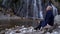 Beautiful middle-aged woman is sitting near waterfall and admiring calm nature at cold day