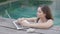 A beautiful mixed-race programmer girl in a blue swimsuit is working on a laptop while standing in the pool. A female