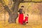 Beautiful mother in a long dress is swinging on a hinged swing little daughter in a red coat in an autumn garden near an old gnarl