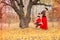 Beautiful mother in a long dress is swinging on a hinged swing little daughter in a red coat in an autumn garden near an old gnarl