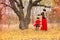 Beautiful mother in a long dress is swinging on a hinged swing little daughter in a red coat in an autumn garden near an old gnarl