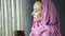 Beautiful muslim arabian woman in pink hijab drinks cofee in front of window