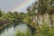 Beautiful nature landscape view of rain drops on pond surface. Rocky coast and tall pine trees on pale sky with rainbow background