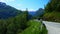 Beautiful panning shot of mountains with snow and forest and a paved road with a guardrail and a car going around the bend.