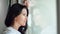 Beautiful pensive brunette female in white shirt looking out of window close-up