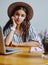 beautiful pensive hipster girl in hat looking at camera working on freelance via netbook