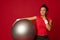 Beautiful pensive multi-ethnic athletic woman holding a fit ball and looking thoughtfully aside isolated over red colored