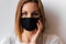 Beautiful portrait of a young woman in a black medical mask and blue eyes and white T-shirt on white background, coquettishly hold