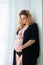 A beautiful pregnant woman in a black robe stands near a bright window. Feminine sexuality, femininity and beautiful pregnancy