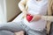 Beautiful pregnant woman sitting on bed and making hand heart on red heart ball at home. Pregnancy, motherhood, love and