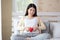 Beautiful pregnant woman sitting on bed and making hand heart on red heart ball at home. Pregnancy, motherhood, love and