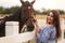 Beautiful pregnant woman stand near the horse. Lady in knitted hat and blue dress. Beautiful farm animal. Brown Hors