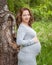 Beautiful pregnant woman well dressed standing near the tree in the green park in sunny day